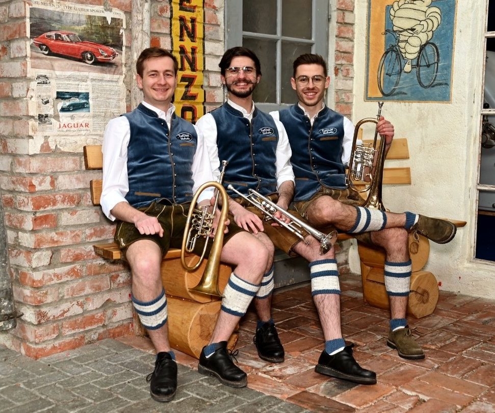 Registerfoto hohes Blech Zwei Flügelhornisten und ein Trompeter sitzen in Tracht und mit ihren Instrumenten auf einer Holzbank und lachen in die Kamera.