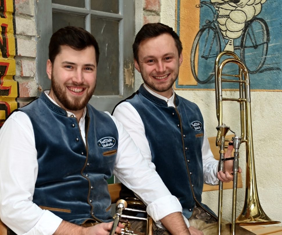 Registerfoto Posaunen Zwei Posaunisten sitzen in Tracht und mit ihren Instrumenten auf einer Holzbank und lachen in die Kamera.