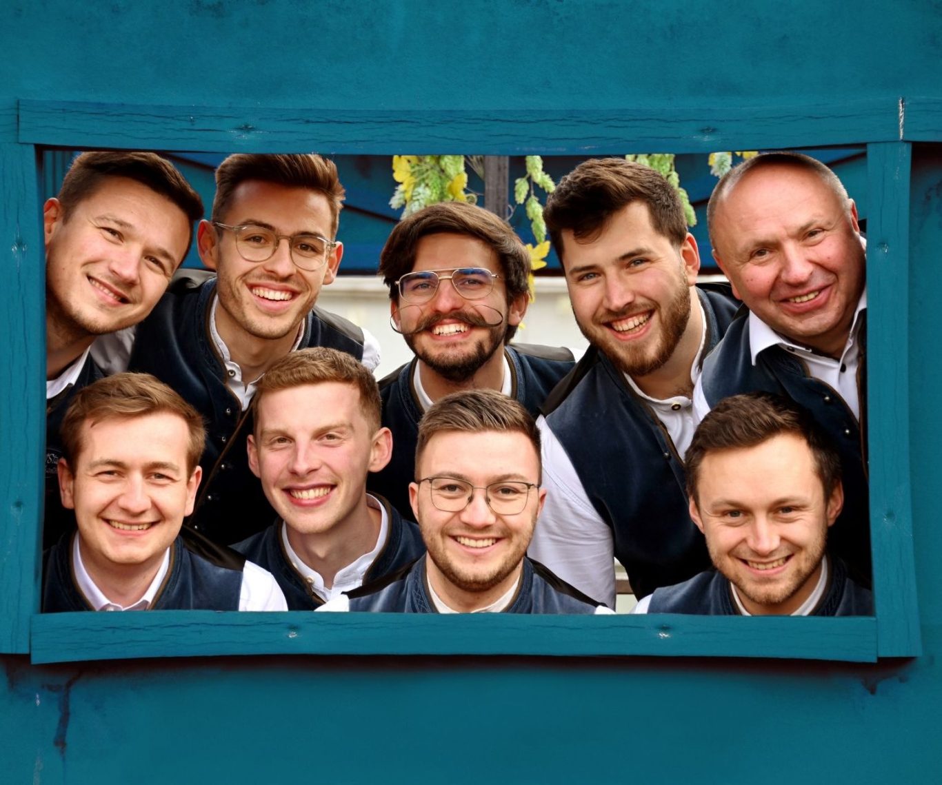Gruppenbild der Feststadelmusikanten Die neun Musiker schauen durch ein Rahmen in einer blauen Holzkiste, man sieht nur ihre lachenden Gesichter.