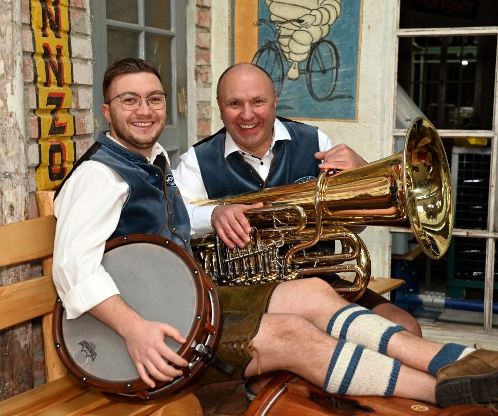 Registerfoto Rhythmusgruppe Ein Schlagzeuger und ein Tubist sitzen in Tracht und mit ihren Instrumenten auf einer Holzbank und lachen in die Kamera.