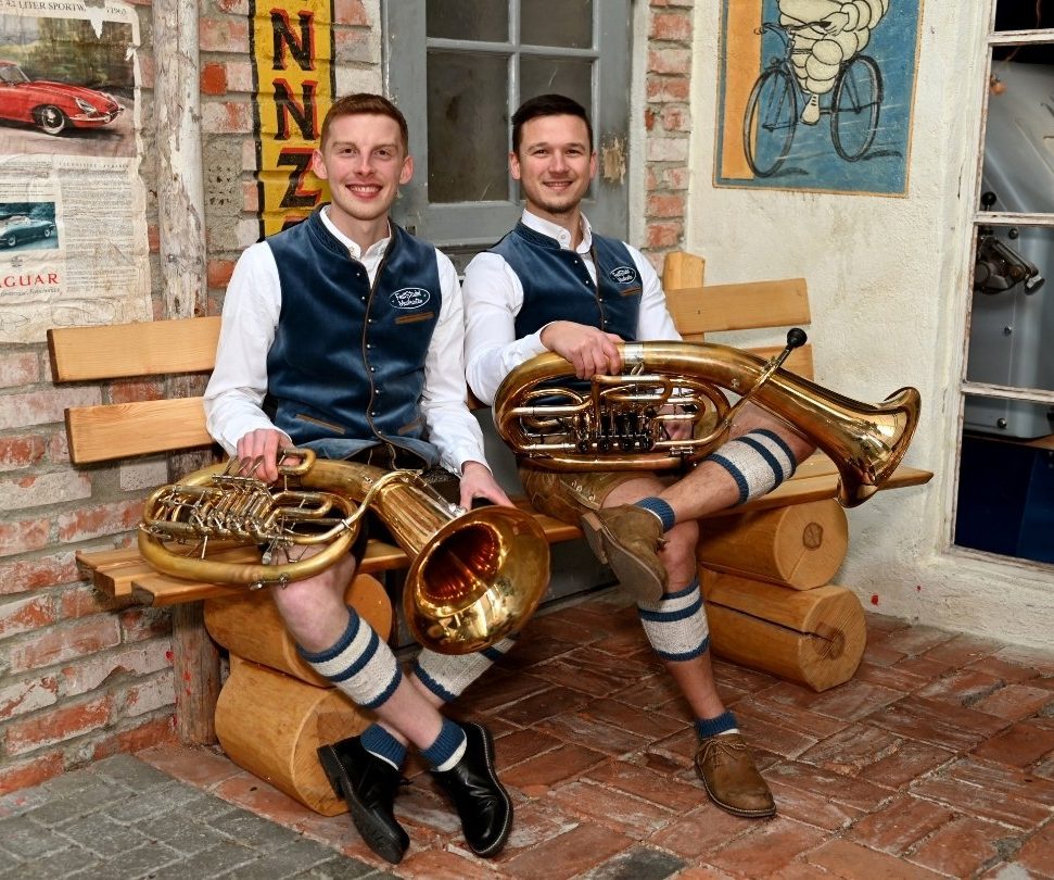 Registerfoto Tenorhorn / Bariton Zwei Tenoristen sitzen in Tracht und mit ihren Instrumenten auf einer Holzbank und lachen in die Kamera.